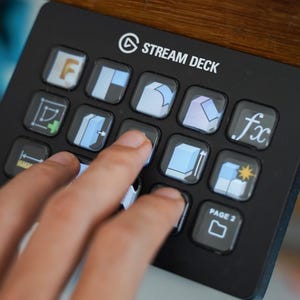 Può includere: Un Stream Deck nero con più pulsanti illuminati, alcuni con icone e testo. Lo Stream Deck è azionato da una mano, con le dita che premono su diversi pulsanti. Lo Stream Deck ha il testo "Stream Deck" in alto.