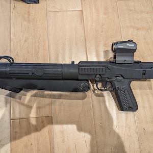 May include: A black blaster prop featuring a long cylindrical barrel with ribbed detailing, a textured pistol grip, and a Sig Sauer red dot optic mounted on the top rail. The device includes a folding lower arm and a rear stock component.