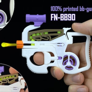 May include: A white 3D printed toy BB gun labeled FN-BB90 with purple accents, a brown internal magazine containing yellow spherical projectiles, and an orange muzzle tip. The device features a top-mounted scope and a skeletal frame design.
