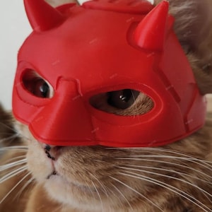 May include: A ginger cat wearing a vibrant red devil mask with horns and eye holes. The mask covers the cat's forehead and eyes. The cat's fur is a mix of orange and white, with long white whiskers.