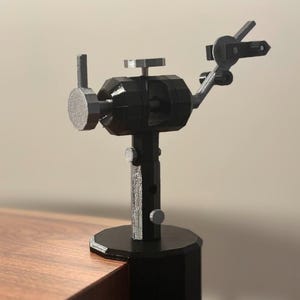 May include: A black and silver articulated hobby stand with a clamp base, a vertical support column with adjustment knobs, and a multi-jointed arm featuring a circular mounting plate and a small clamp mechanism for holding small items securely in place.