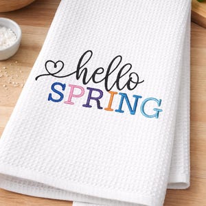 May include: White waffle-weave kitchen towel with the embroidered text "hello SPRING" in black, pink, purple, orange, and blue. The towel is folded and rests on a wooden surface, suggesting a spring or kitchen theme.