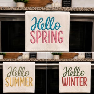 Op de afbeelding: Een set van drie off-white keukendoeken met geborduurde tekst. Eén doek toont Hello SPRING in blauw en roze, één toont Hello SUMMER in groen en geel, en één toont Hello WINTER in groen en rood, in een combinatie van script- en blokletters.