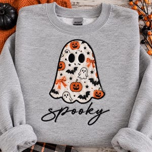 May include: A heather gray crewneck sweatshirt featuring a large central graphic of a ghost shape filled with a pattern of orange pumpkins, black bats, small white ghosts, and orange bows. The word spooky is written below in black script.