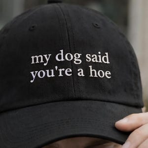 Pode incluir: Boné de beisebol preto com o texto bordado "my dog said you're a hoe" em branco. O boné tem uma aba curva e um botão superior. Um acessório casual.