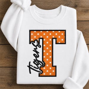 May include: A white long-sleeve sweatshirt featuring a large, orange, polka-dot letter T with a black embroidered outline. The word Tigers is written in black cursive script across the left side of the letter T, creating a bold graphic design.