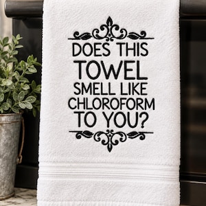 May include: A white hand towel featuring black embroidered text that reads DOES THIS TOWEL SMELL LIKE CHLOROFORM TO YOU? The text is framed by decorative black scrollwork at the top and bottom, and the towel has a textured border near the bottom edge.