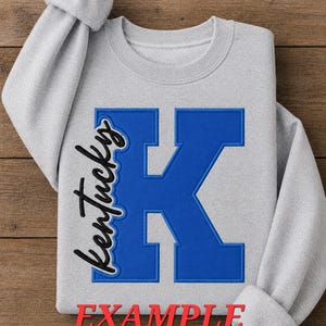 May include: A light gray crewneck sweatshirt featuring a large, bold blue letter K embroidered on the front. The word Kentucky is embroidered in black cursive script along the left side of the letter. The red text EXAMPLE is printed at the bottom.
