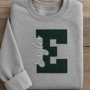 Puede incluir: Una sudadera gris jaspeado de cuello redondo con una letra E mayúscula de bloque en color verde oscuro bordada en el centro del pecho. La palabra Eagles está cosida en letra cursiva blanca sobre el lado izquierdo de la letra E.