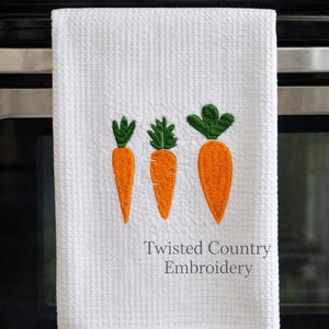 Könnte beinhalten: Weißes Küchenhandtuch mit Waffelstruktur und drei gestickten orangefarbenen Karotten und grünen Blättern. Der Text "Twisted Country Embroidery" ist darunter gedruckt. Das Handtuch hängt an einem Edelstahlgerät.