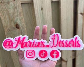 May include: A white acrylic sign featuring the bright pink text @Maria's.Desserts in a cursive font, positioned above three circular icons representing Instagram, TikTok, and Facebook, all rendered in a matching vibrant pink color.