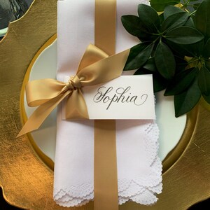May include: A white cloth napkin with scalloped edges wrapped in a gold satin ribbon tied in a bow. A rectangular white place card with the name Sophia written in black cursive script is tucked under the ribbon. A gold charger plate sits below.