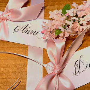 May include: Two white rectangular gift tags featuring the names Anne and Diego written in black cursive script. Each tag is adorned with a light pink satin ribbon tied into a bow. A cluster of small, light pink artificial flowers is positioned nearby.
