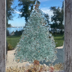 May include: A framed glass art piece depicting a Christmas tree made of green glass shards with a gold star on top. The tree is surrounded by white seashells and sand.