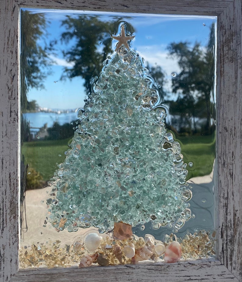 May include: A framed glass art piece depicting a Christmas tree made of green glass shards with a gold star on top. The tree is surrounded by white seashells and sand.