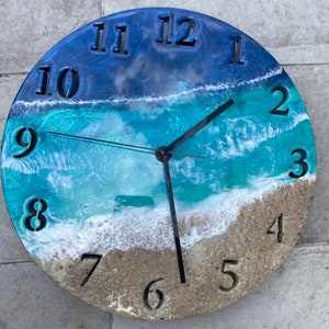 May include: A round clock with a blue and green ocean wave design. The clock face has black Roman numeral hour markers and black hands. The clock is made of resin and has a white sandy beach design at the bottom.
