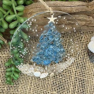 May include: A clear, round Christmas ornament with a blue, textured tree design. The tree is topped with a white star and sits on a bed of sand and seashells. A twine loop is attached for hanging.