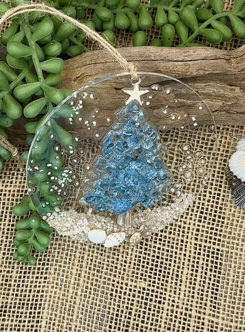 May include: A clear, round Christmas ornament with a blue, textured tree design. The tree is topped with a white star and sits on a bed of sand and seashells. A twine loop is attached for hanging.