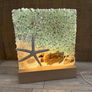 May include: A decorative lamp featuring a starfish, seashells, and a light-up resin panel. The panel has a green and clear textured design, with a wooden base. The lamp is designed to resemble a beach scene.