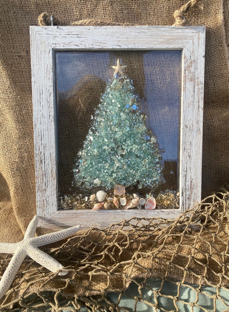 May include: A white framed wall hanging featuring a Christmas tree crafted from blue and green glass pieces. The tree is adorned with a gold star and surrounded by seashells.