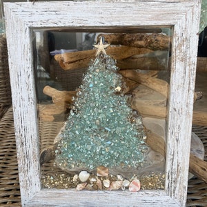 May include: A whitewashed wooden frame with a glass front, displaying a Christmas tree crafted from blue and green glass pieces. The tree is adorned with a gold star and surrounded by seashells and gold glitter.