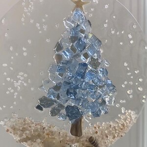 May include: A clear, circular Christmas ornament featuring a blue glass tree, a starfish topper, and sand at the base. Small seashells are nestled in the sand. The ornament is suspended by a natural twine loop.