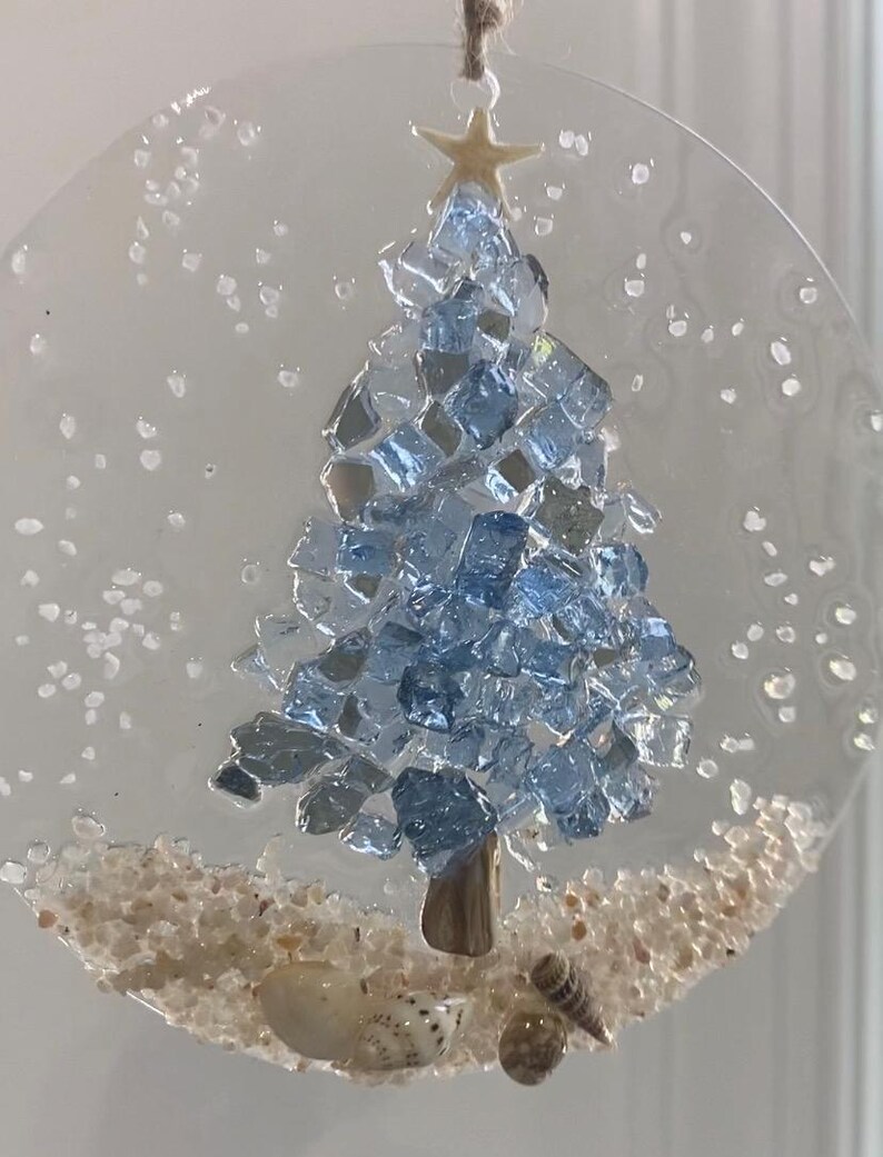 May include: A clear, circular Christmas ornament featuring a blue glass tree, a starfish topper, and sand at the base. Small seashells are nestled in the sand. The ornament is suspended by a natural twine loop.