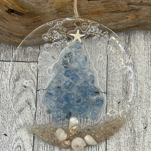 May include: Clear glass Christmas ornament with a blue tree design made of small, square blue stones. The ornament is decorated with sand, seashells, and a starfish. A natural twine loop is attached for hanging.