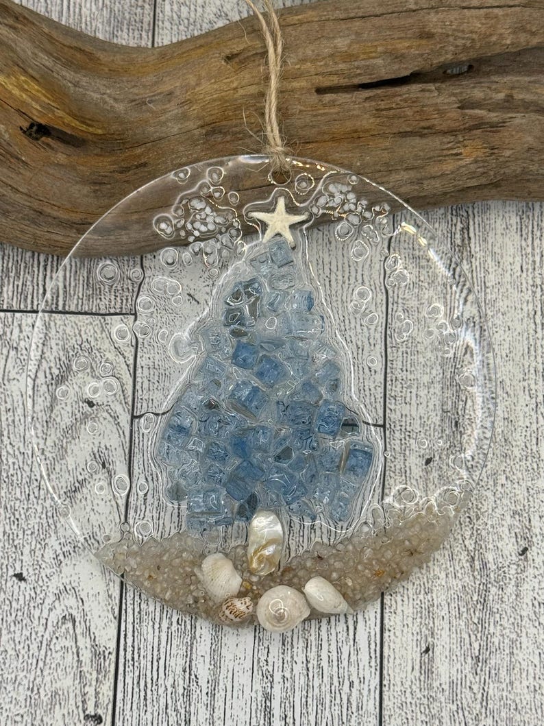 May include: Clear glass Christmas ornament with a blue tree design made of small, square blue stones. The ornament is decorated with sand, seashells, and a starfish. A natural twine loop is attached for hanging.