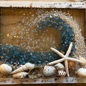 May include: A framed beach-themed art piece. The artwork features a wave design made of blue glass, sand, and seashells. A white starfish and various seashells are arranged at the bottom. The frame is white with a distressed finish, giving it a rustic look.