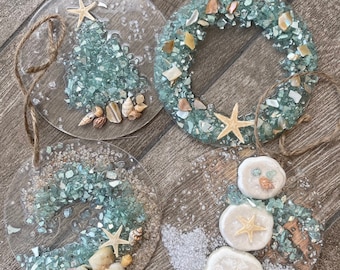 Könnte beinhalten: Vier runde, klare Glaskugeln mit Strandthema. Eine Kugel zeigt einen grünen und blauen Weihnachtsbaum mit Muscheln und einem Seestern. Eine weitere Kugel zeigt einen Kranz aus blauen und grünen Glassplittern mit einem Seestern in der Mitte. Die dritte Kugel zeigt eine blaue und grüne Welle mit Muscheln und einem Seestern. Die vierte Kugel zeigt einen Schneemann aus weißen Muscheln mit einem blauen und grünen Schal und einem Seestern auf jeder der drei Schichten des Schneemanns.