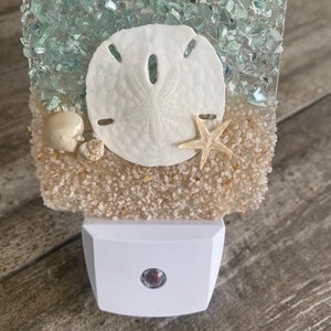 May include: A decorative night light featuring a sand dollar, starfish, and seashells embedded in a clear resin. The top section has blue-green glass pieces, and the base is white. The night light is designed to plug into a wall outlet.