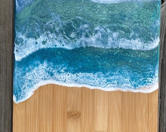 May include: A wooden cutting board with a blue and white epoxy resin design that resembles ocean waves. The resin is clear and has a glossy finish. The wood is a light brown color and has a natural grain.