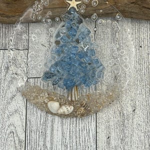 May include: A clear, circular ornament featuring a blue glass Christmas tree design. The tree sits on a bed of sand with seashells. A small starfish hangs from a twine loop at the top.