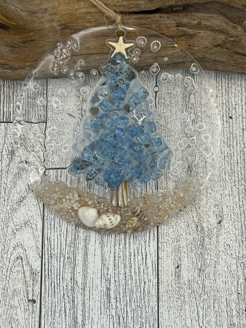 May include: A clear, circular ornament featuring a blue glass Christmas tree design. The tree sits on a bed of sand with seashells. A small starfish hangs from a twine loop at the top.