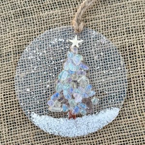 May include: Clear, circular Christmas ornament with a tree made of colorful stones and a star on top. The ornament has a jute string for hanging and a white, snowy base. A festive holiday decoration.