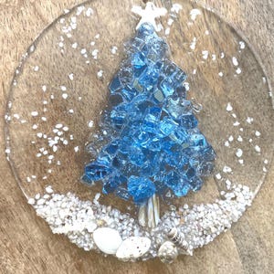 May include: A clear, round Christmas ornament with a blue glass tree design. The tree is surrounded by white sand and small seashells. A white star tops the tree, and a twine loop is attached for hanging.