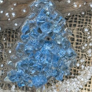 May include: A handcrafted Christmas tree made of blue glass pieces, topped with a white starfish. The tree is set on a bed of sand and seashells, with a burlap background and driftwood accents. A unique coastal holiday decoration.