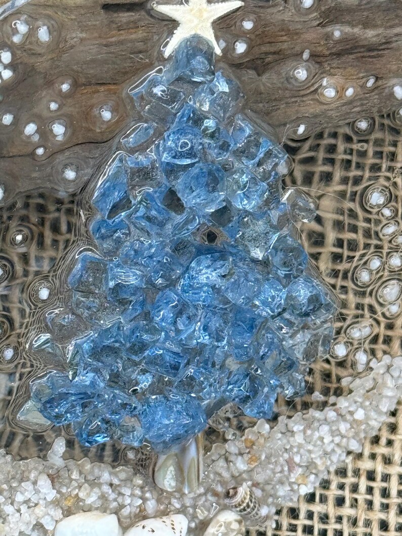 May include: A handcrafted Christmas tree made of blue glass pieces, topped with a white starfish. The tree is set on a bed of sand and seashells, with a burlap background and driftwood accents. A unique coastal holiday decoration.
