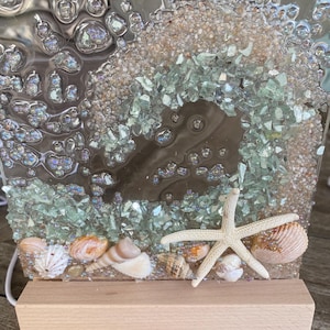 May include: A decorative resin art piece featuring a beach theme. The artwork includes seashells, a starfish, and crushed glass in shades of green and beige. The piece is mounted on a rectangular wooden base.