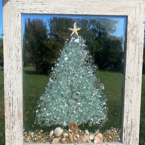 May include: A white framed glass Christmas tree bauble with a blue and green glass mosaic design, a gold star on top, and seashells at the base.