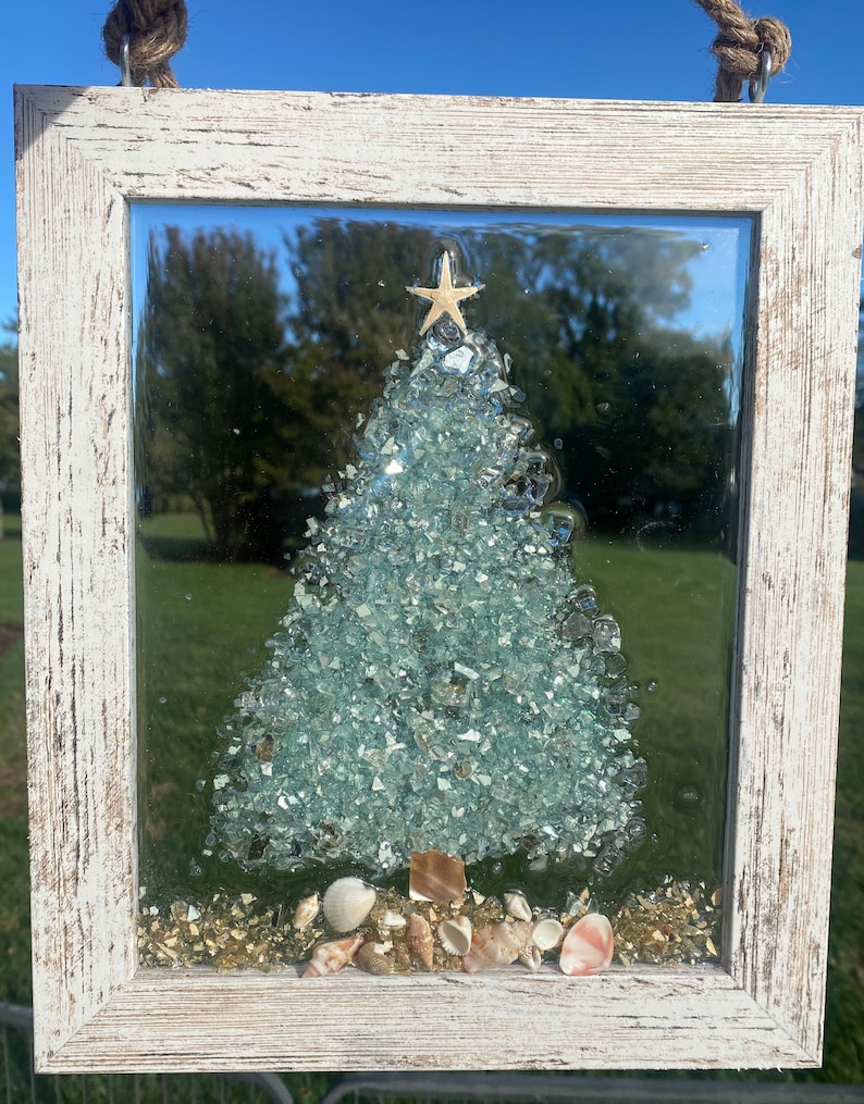 May include: A white framed glass Christmas tree bauble with a blue and green glass mosaic design, a gold star on top, and seashells at the base.