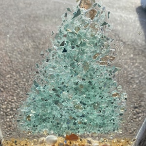 May include: A framed, clear resin artwork featuring a green, glittery Christmas tree with a gold star on top. The tree is surrounded by small, white and brown seashells on a sandy base.
