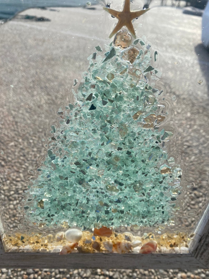 May include: A framed, clear resin artwork featuring a green, glittery Christmas tree with a gold star on top. The tree is surrounded by small, white and brown seashells on a sandy base.