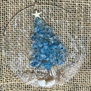 May include: A clear, circular Christmas ornament with a blue, mosaic-style Christmas tree design. A white starfish sits atop the tree. The base of the tree is adorned with seashells and white sand. A natural twine loop is at the top.