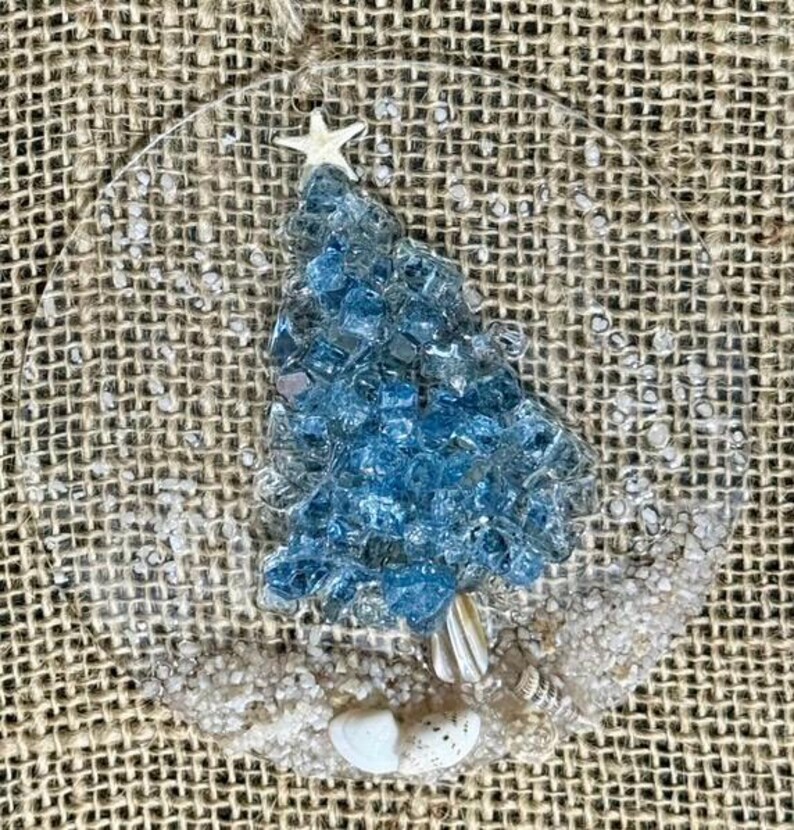 May include: A clear, circular Christmas ornament with a blue, mosaic-style Christmas tree design. A white starfish sits atop the tree. The base of the tree is adorned with seashells and white sand. A natural twine loop is at the top.