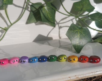 May include: A row of colorful, handcrafted clay beads, each with a painted face, arranged in a rainbow spectrum. The beads are in shades of pink, purple, blue, green, yellow, and red. The background includes green leaves.
