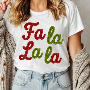 Könnte beinhalten: Weißes T-Shirt mit dem Text "Fa la la" in roter und grüner, seilartiger Schrift. Das Shirt ist ein Rundhalsausschnitt und wird mit blauen Jeans und einer hellen Strickjacke getragen.