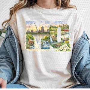 May include: Cream-colored t-shirt featuring the letters "MO" with watercolor illustrations. The "M" showcases a river scene with a waterfall and a cityscape, while the "O" displays a rural landscape with a red building and flowers.