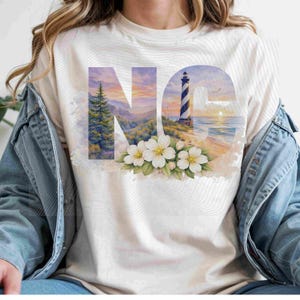 May include: Cream-colored t-shirt featuring a watercolor design of a lighthouse, ocean, and sunset within the letters "NC". White flowers and green leaves are at the bottom of the design. The shirt is made of a soft material.